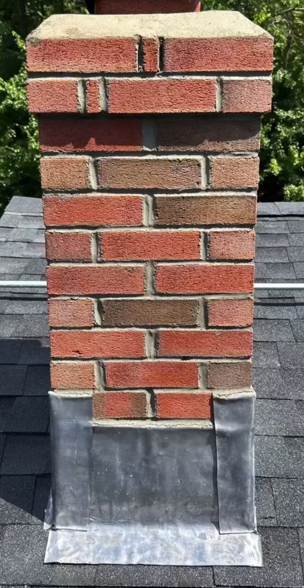 Brick chimney extending from a gray roof, with metal flashing around the base.