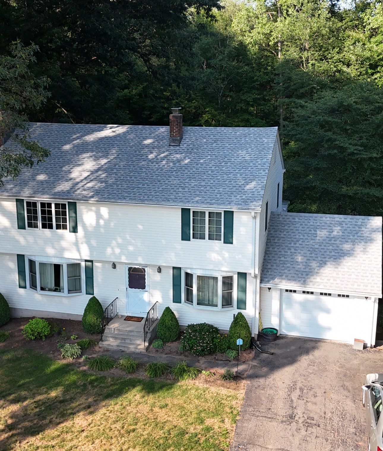 Two-story white house with gray roof, green shutters, and attached garage; surrounded by trees.