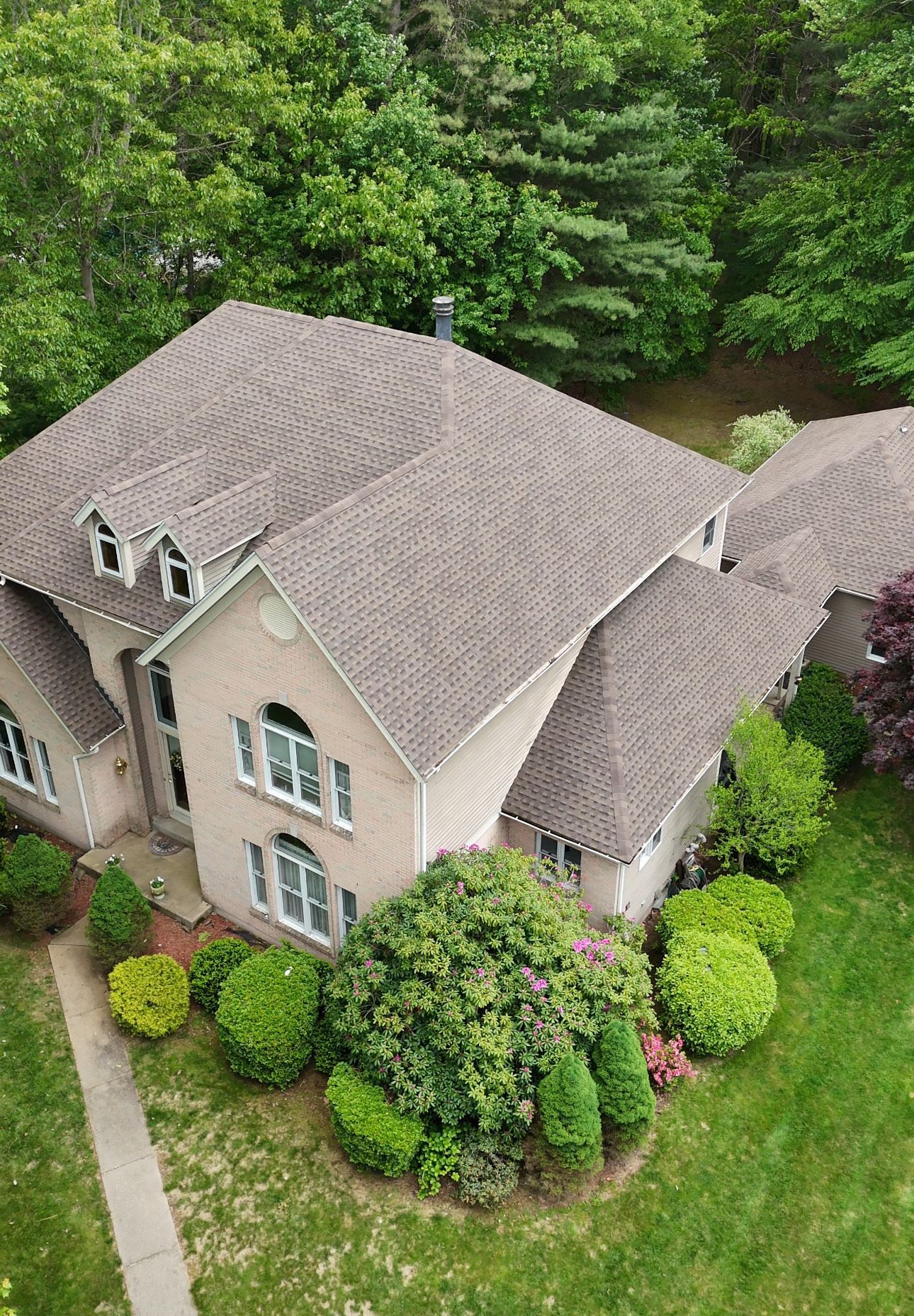 Aerial view of a beige house with a brown roof and surrounding green trees and grass.