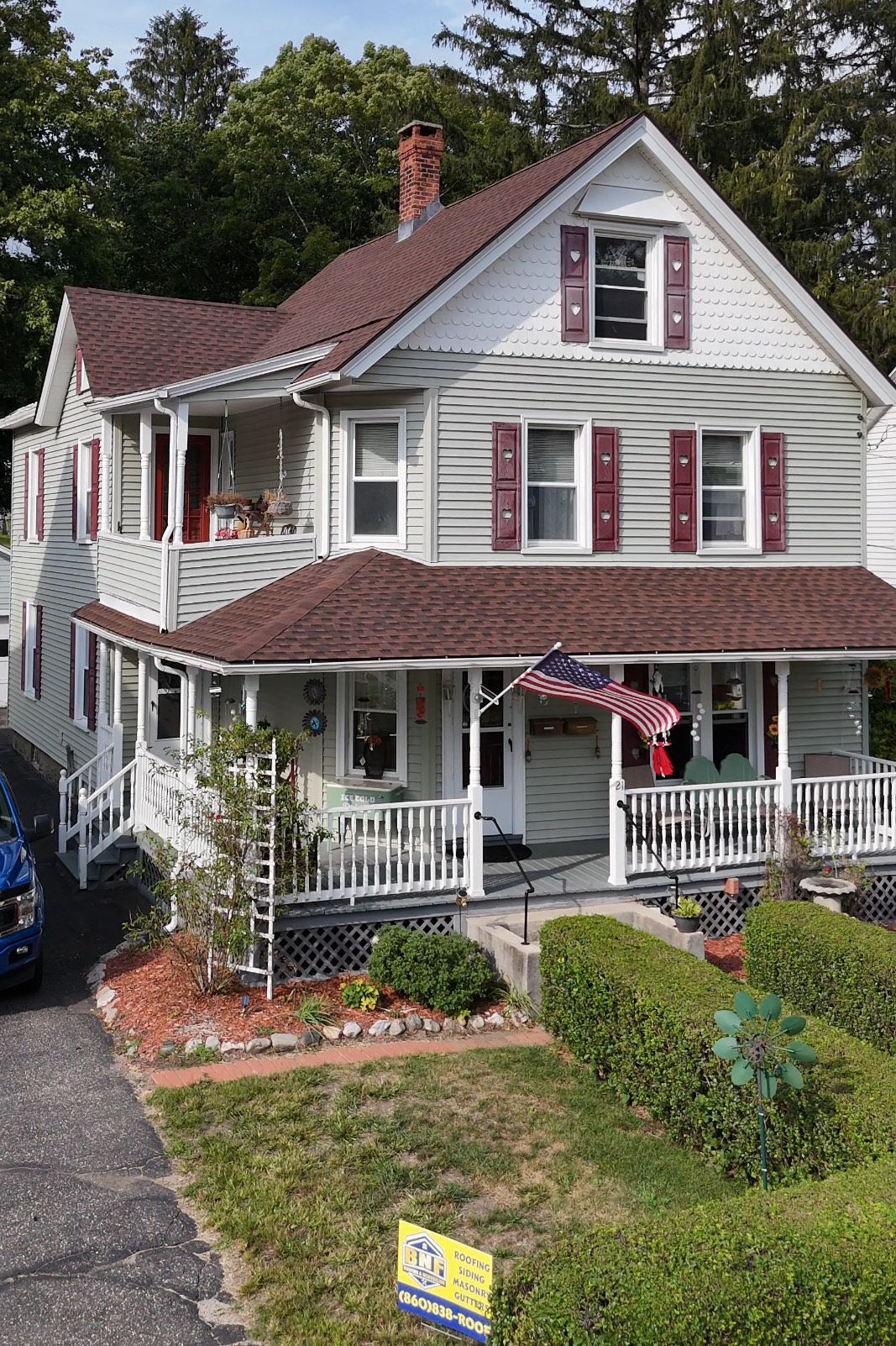 Two-story house with light siding, red shutters, and porch. American flag hangs from the porch, and there's a small garden.