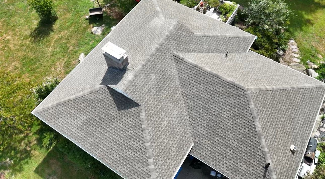 Aerial view of a gray shingled roof with a chimney, surrounded by green grass and trees.