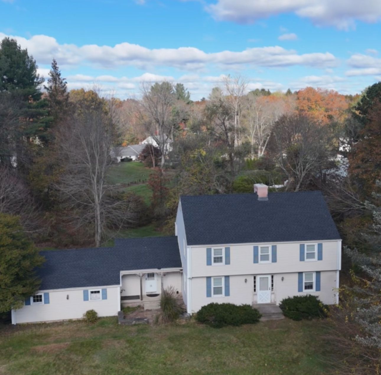 Aerial view of a two-story white house with a black roof and blue shutters, surrounded by autumn trees under a cloudy sky.