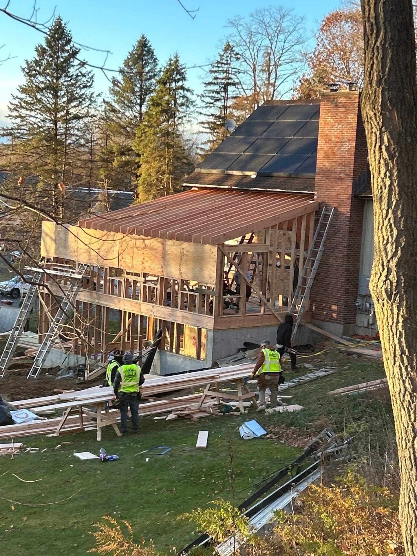 Construction workers build an addition onto a brick house. One worker gestures, wearing a safety vest.