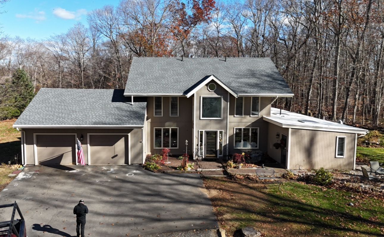 Two-story house with gray roof, attached garage, and a long driveway. Man stands near the driveway.