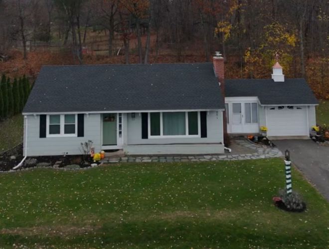 Gray ranch-style house with dark roof and white trim, set on a green lawn with trees in the background.