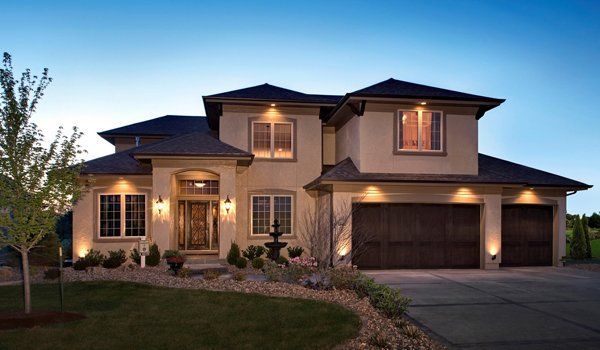 Two-story beige house with dark roof, brown garage doors, lit by outdoor lighting, and landscaped yard.