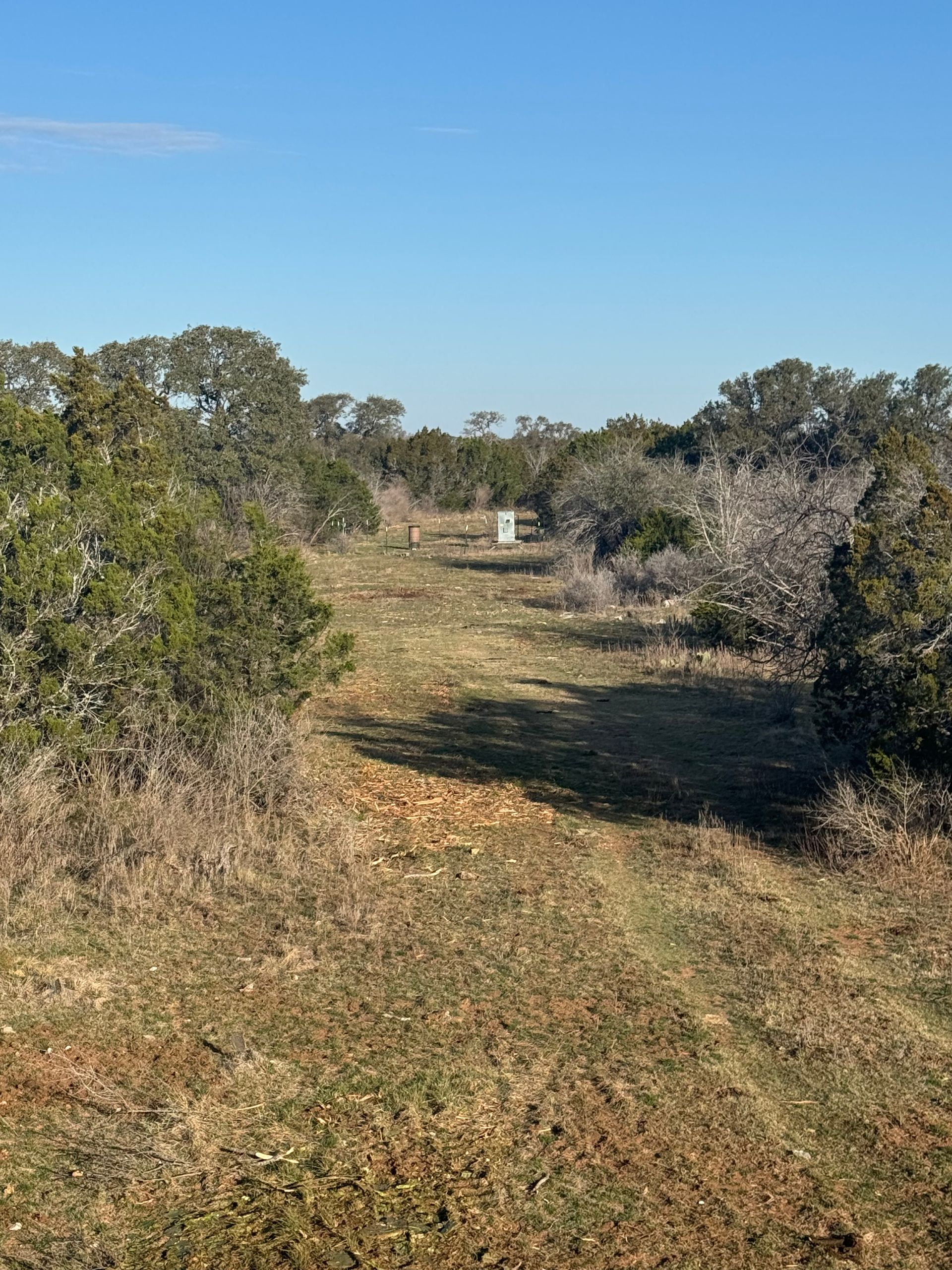 A path through a field lined with trees, leading to a white box under a blue sky.