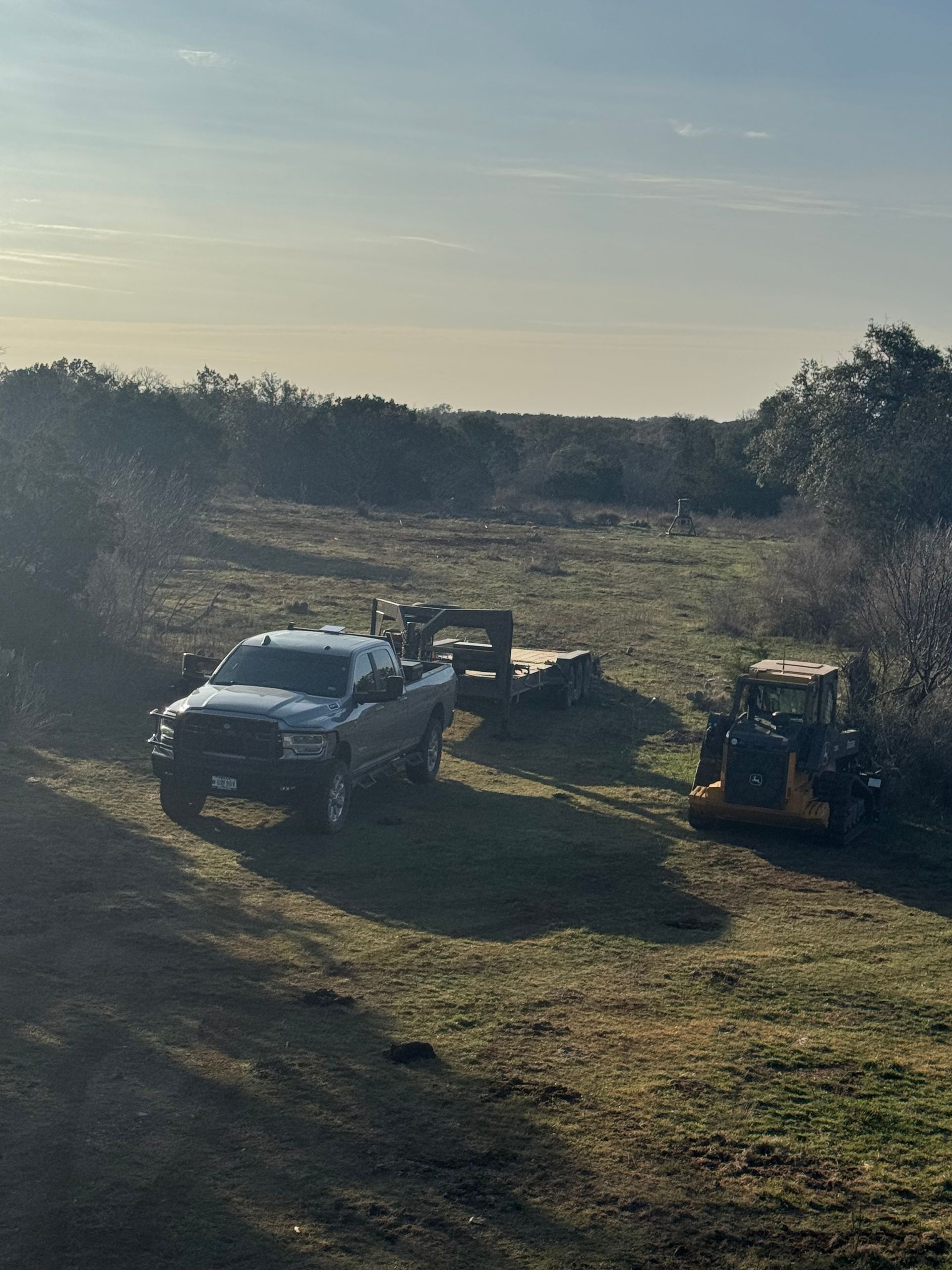 Truck with a trailer and small bulldozer in a grassy field surrounded by trees under a sunny sky.