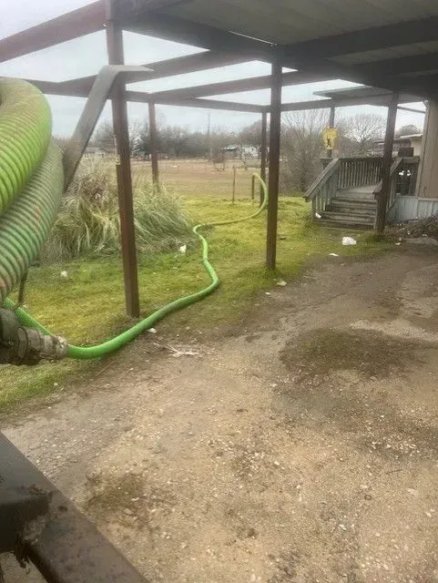 Green hose runs across a grassy yard, under a brown-framed shelter.