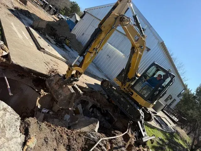 Yellow excavator demolishing concrete next to a building. Operator visible in cab.