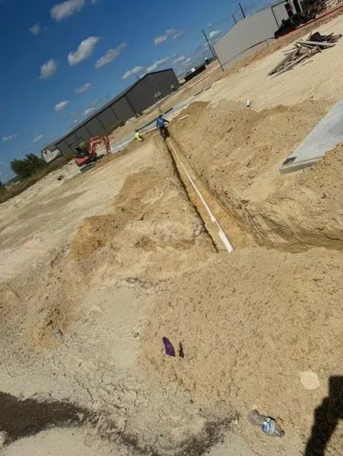 A trench in sandy soil, with white pipe visible, being worked on at a construction site on a sunny day.