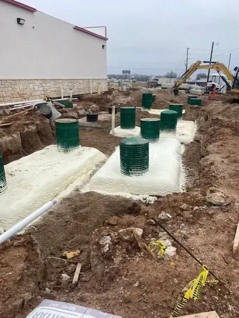 Construction site: foam-insulated foundation with green wire mesh columns, trench, and excavator in background.