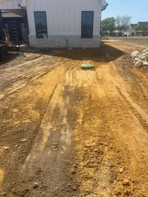 Dirt construction site; house in background.  Yellow dirt road with tire tracks; green septic lid in the middle.