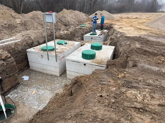 Construction site with three concrete septic tanks in a trench. Two men stand nearby.