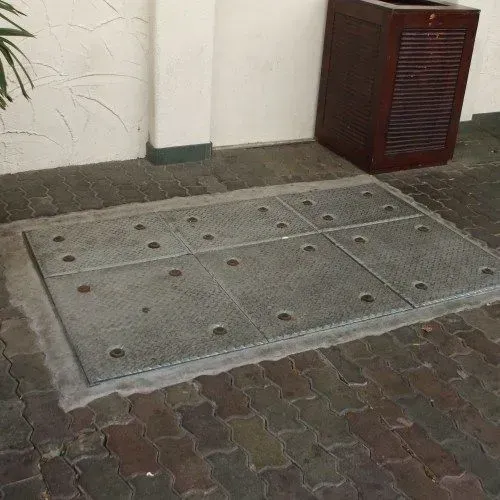 Gray metal grate on a brick patio, near a brown wooden box and a white wall.