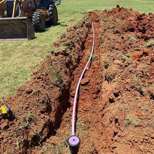 A long, narrow trench in red dirt with a purple pipe, next to a tractor on a grassy field.