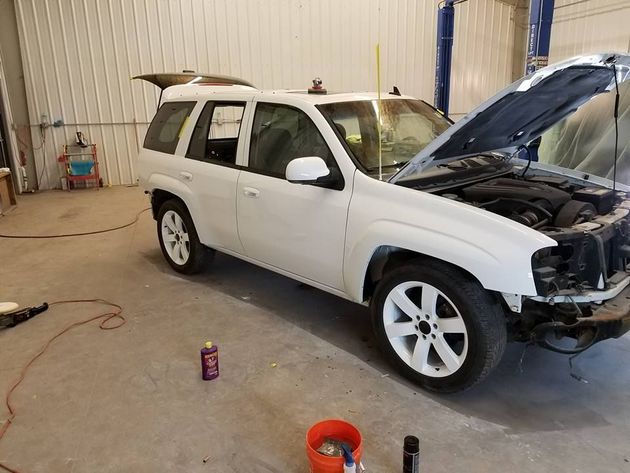 White SUV in a garage with front end removed, hood open, and tools on the floor.