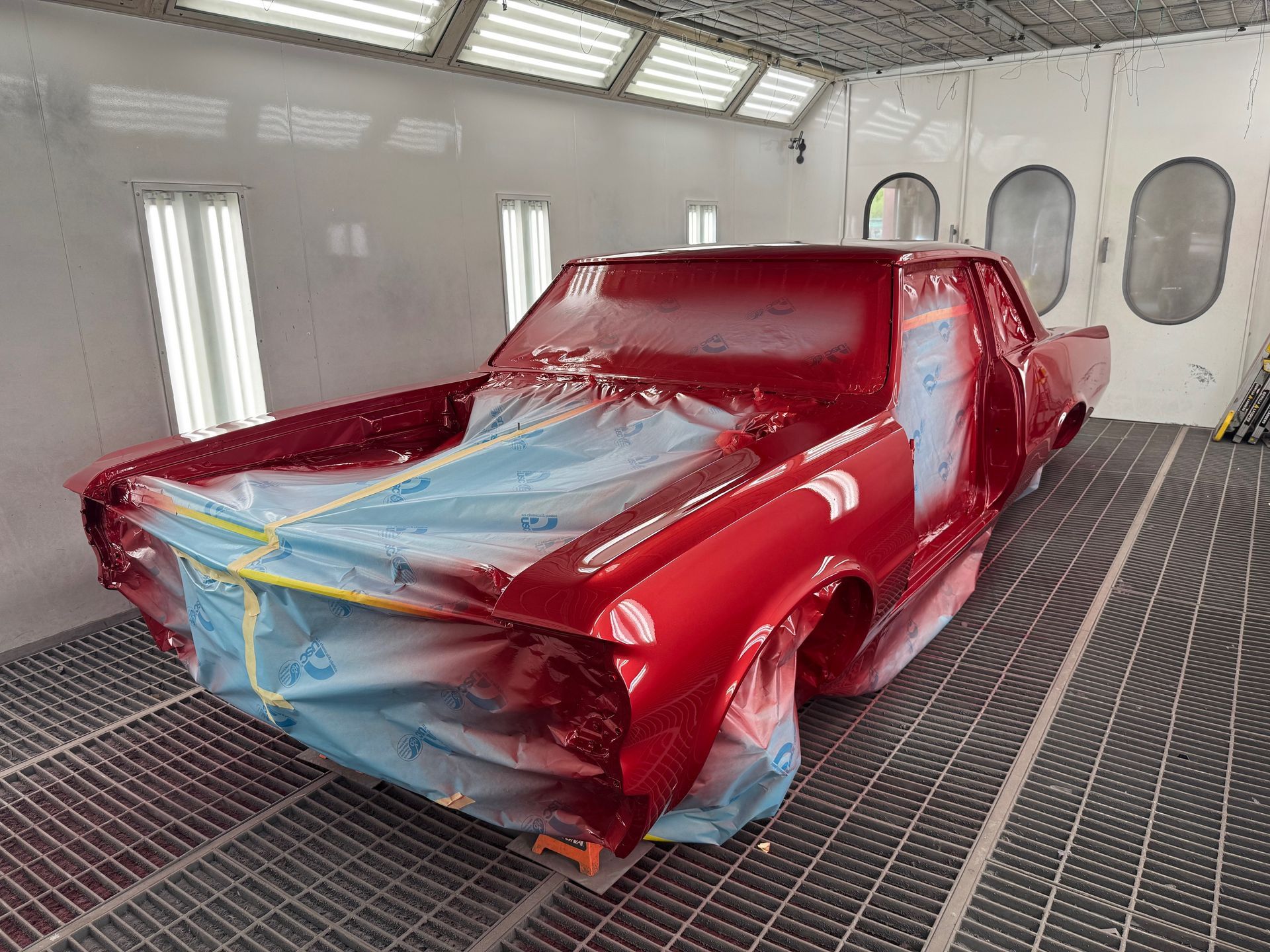 Red car body in a paint booth, masked with blue tape and plastic sheeting.