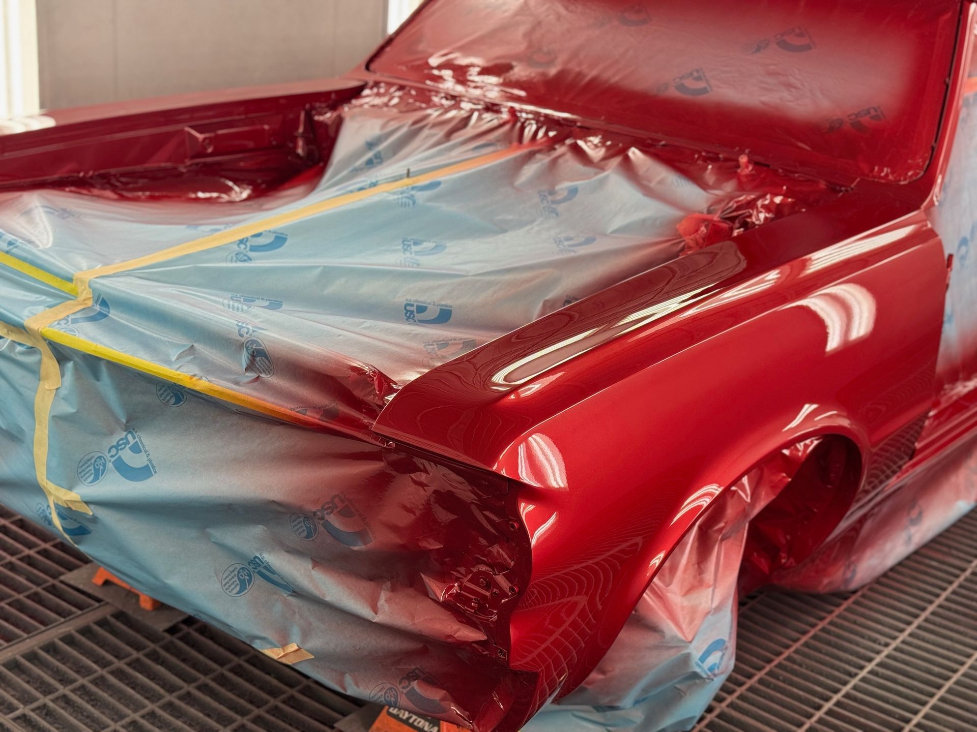 Red car body wrapped in blue masking plastic and tape in a paint booth