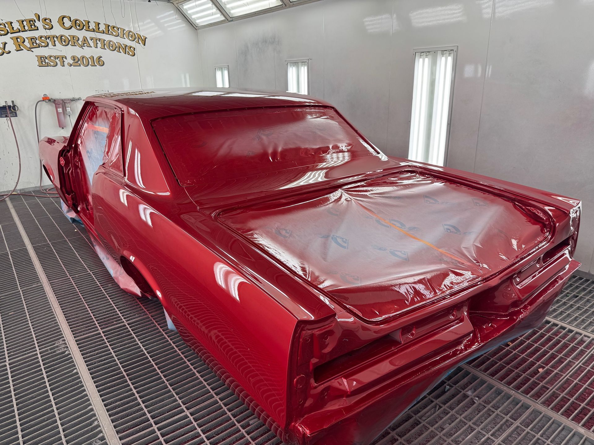 Red car body in a paint booth, covered in glossy red primer with silver hood areas.