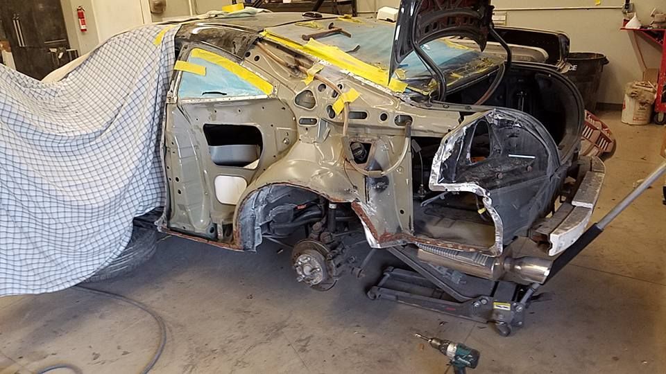 Severely damaged car body in a garage, with exposed metal and detached parts.