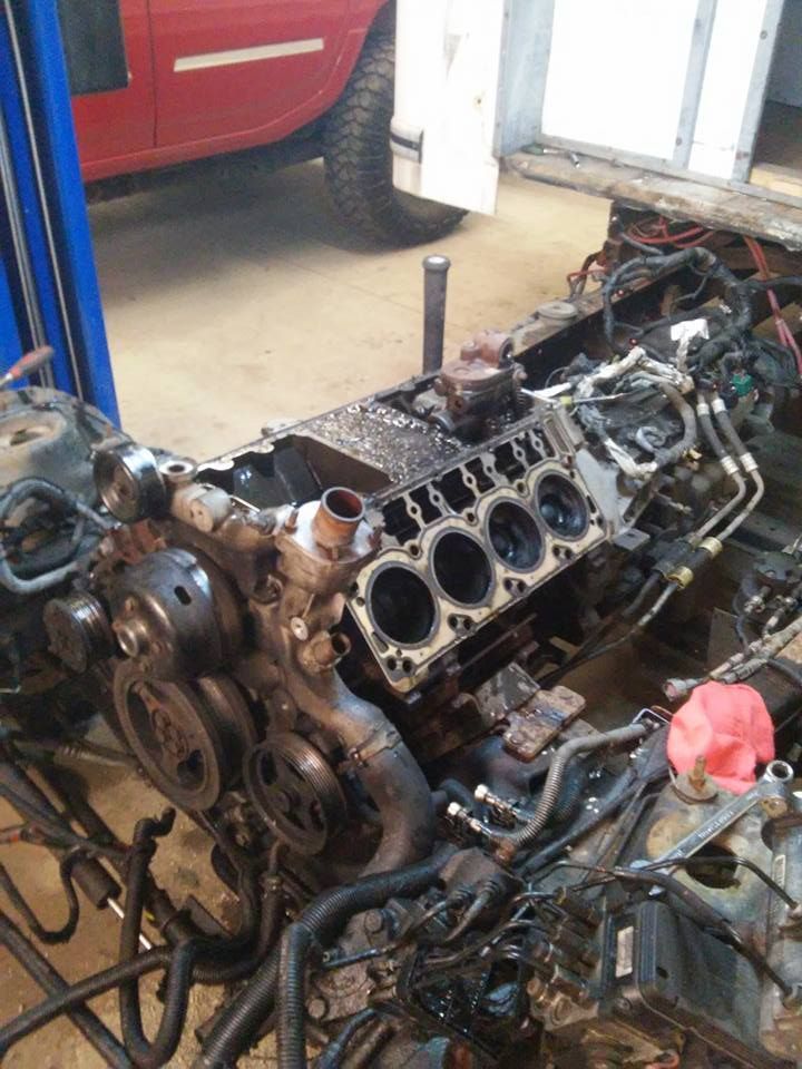 Car engine block on a workbench in a garage, surrounded by hoses and parts, with a red car in the background