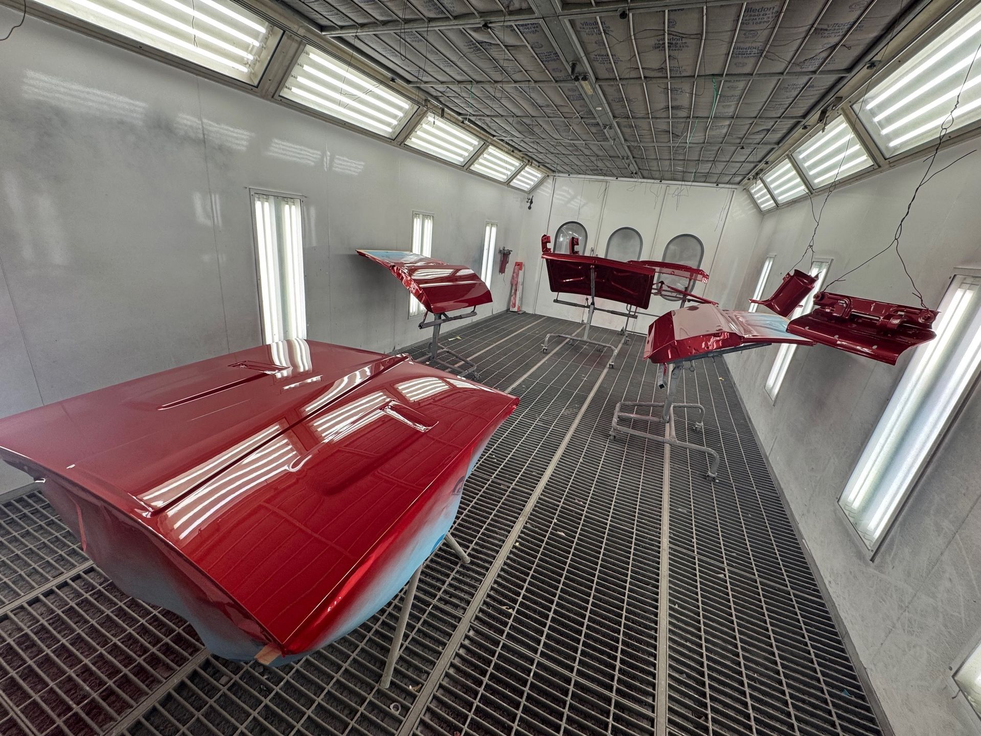 Red-painted aircraft parts in a bright industrial paint booth with grated floor and overhead lights