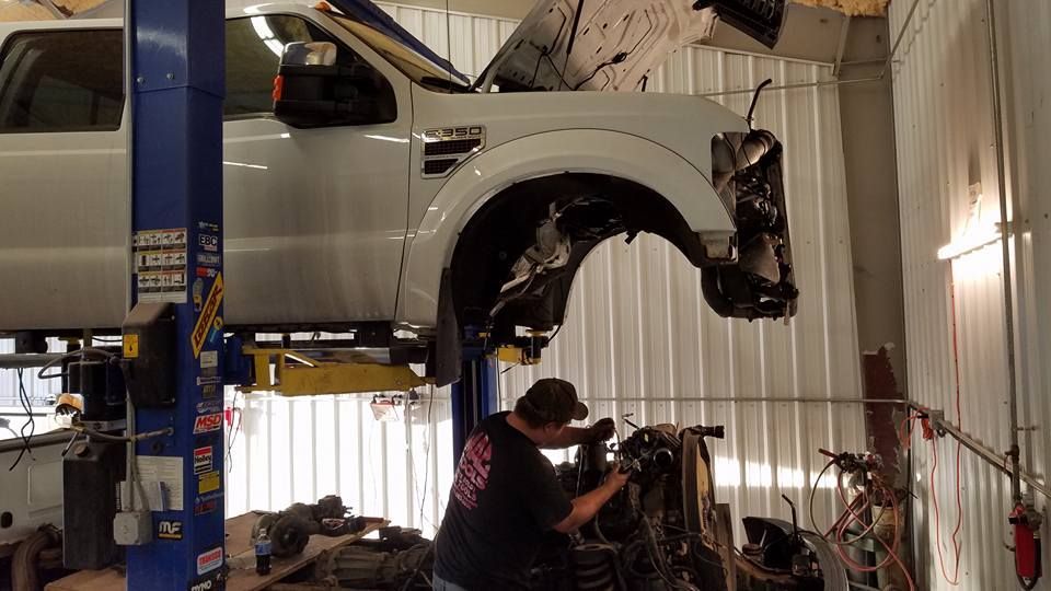 White pickup truck raised on a lift in an auto repair shop, with mechanics working underneath.