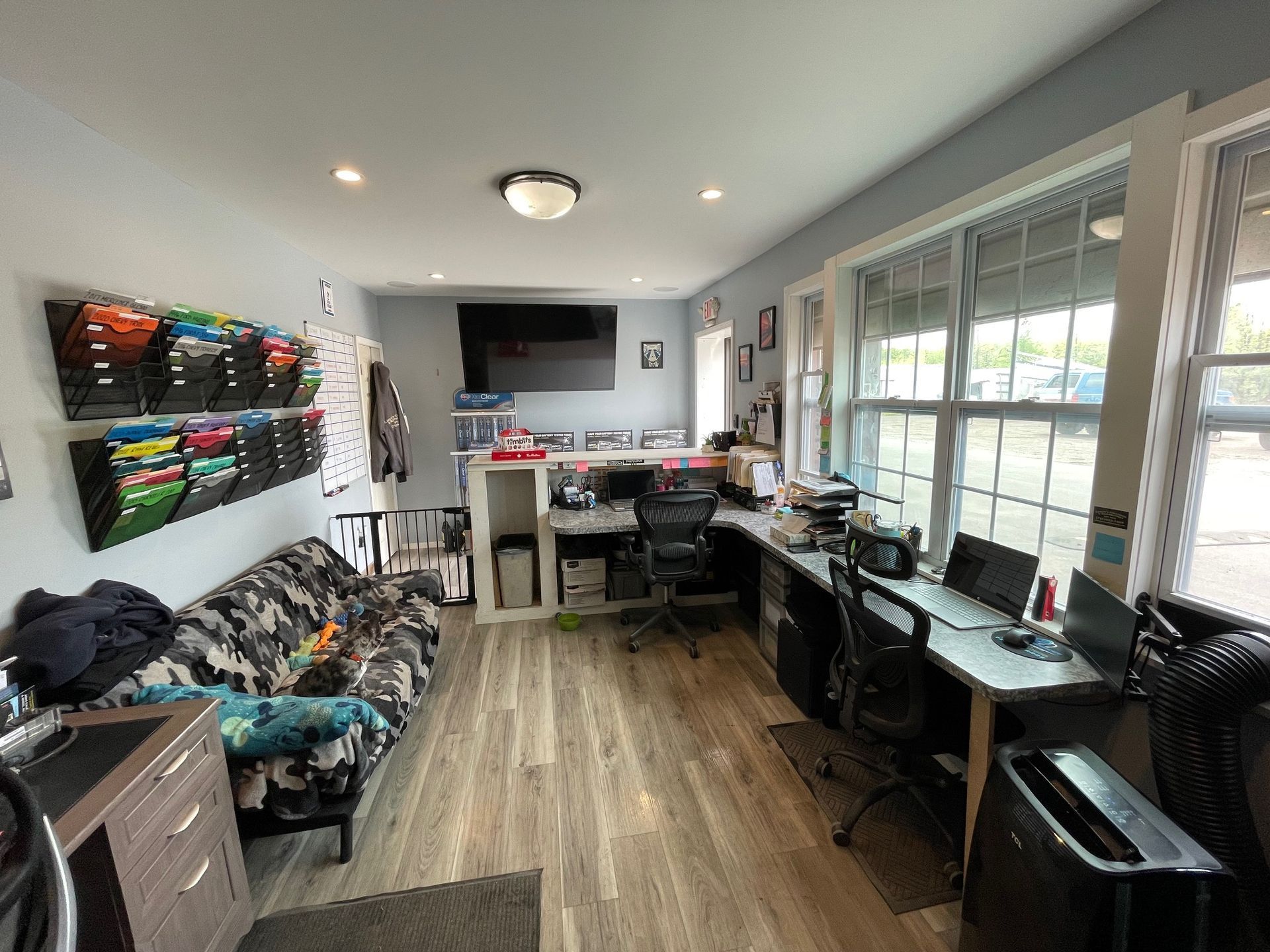 Cluttered home office with desks, computer monitors, wall art, and a couch by large windows