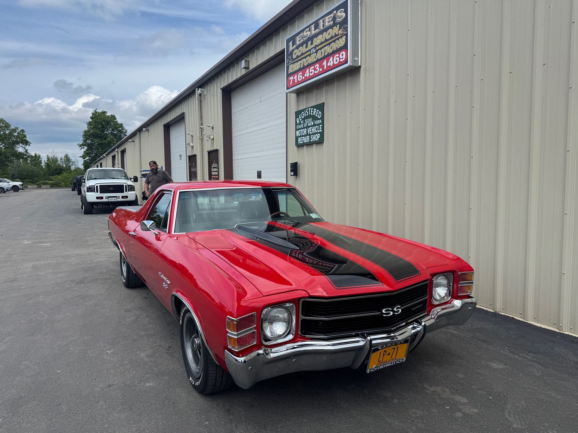 Red classic car with black racing stripes parked beside an industrial building.