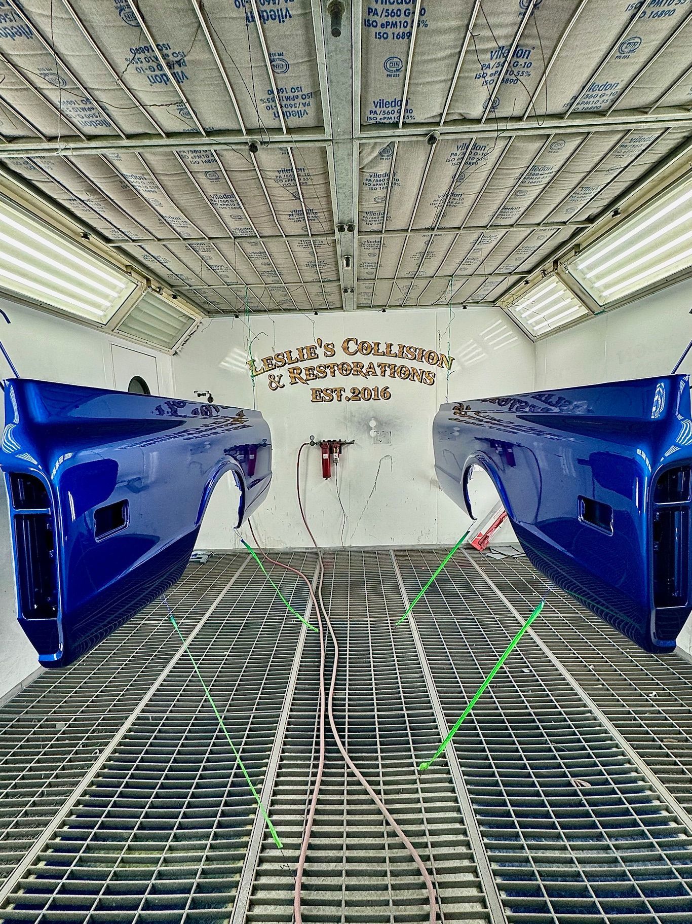 Blue car body panels inside an industrial spray booth with gridded floor and overhead vents