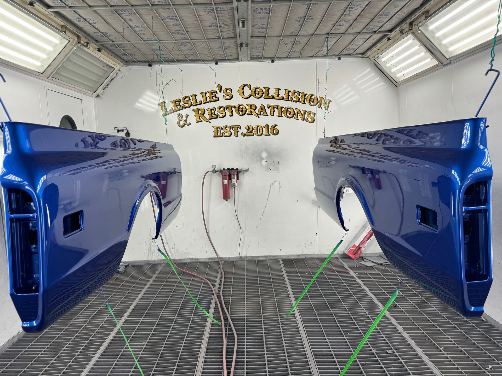Blue car body panels in a spray booth, with a white wall and gold sign in the background.