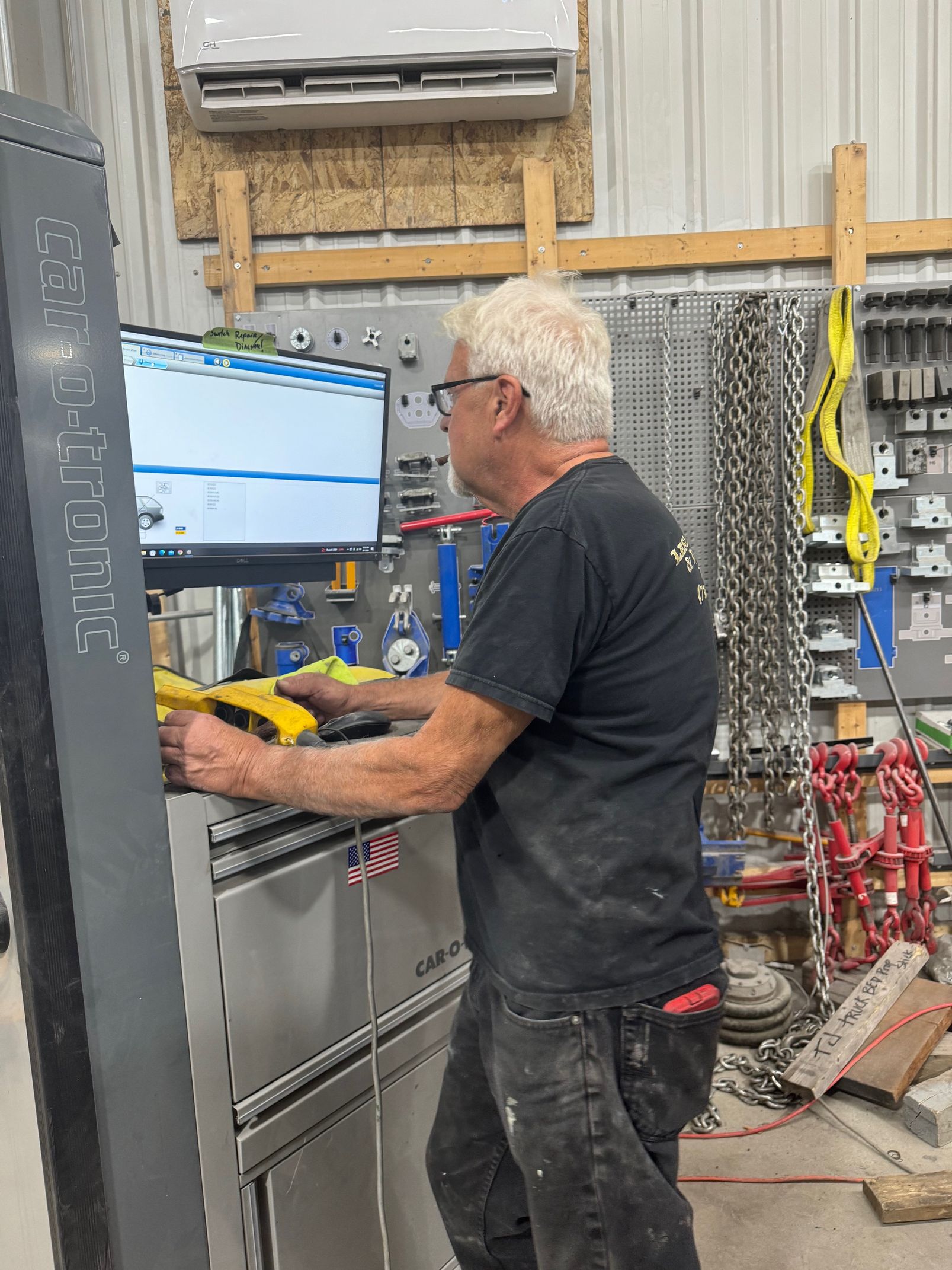 Technician at a computer in an industrial workshop, using tools beside wall-mounted equipment.