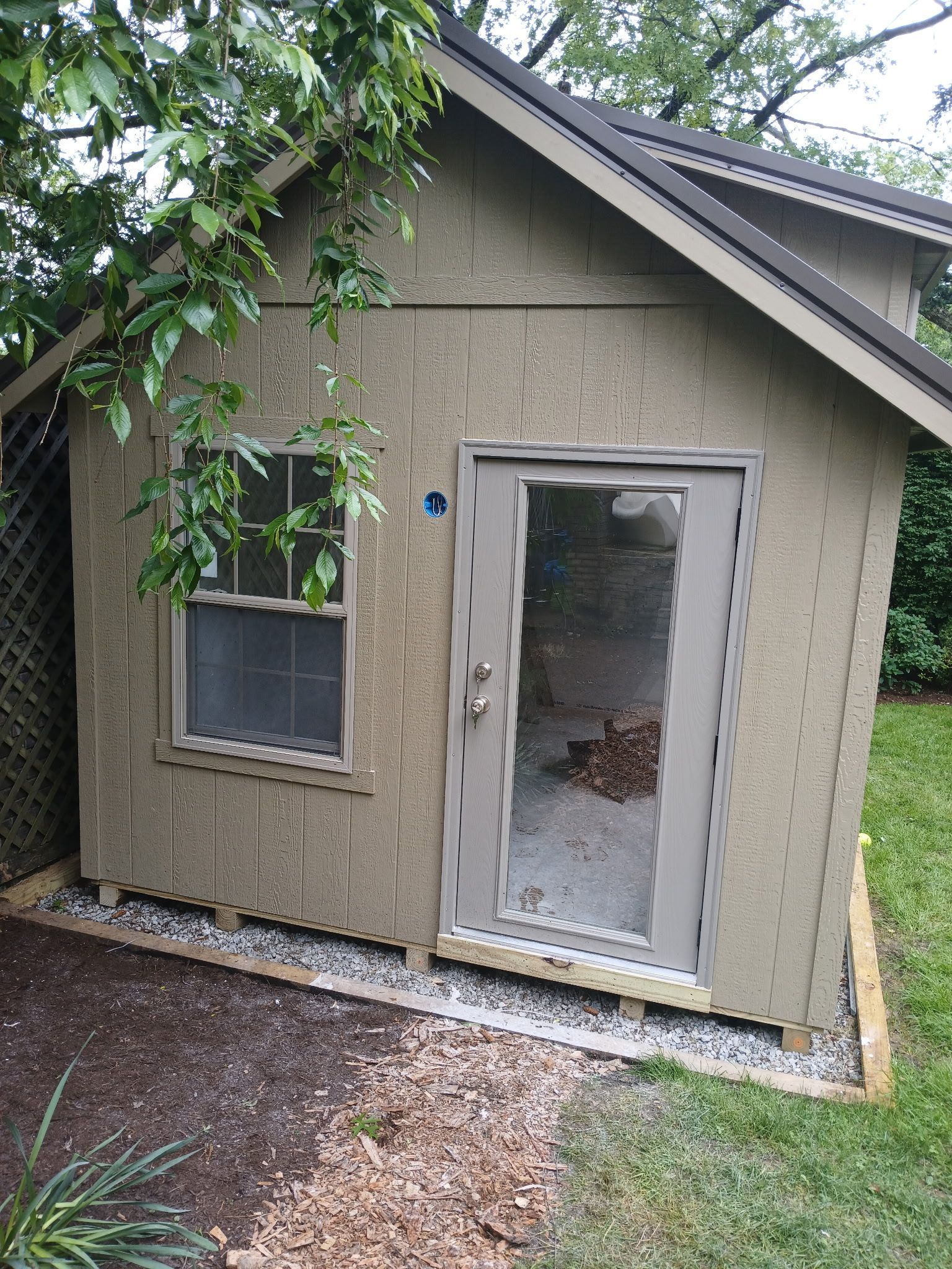 Small tan shed with a white door and window, viewed from the front under tree branches.
