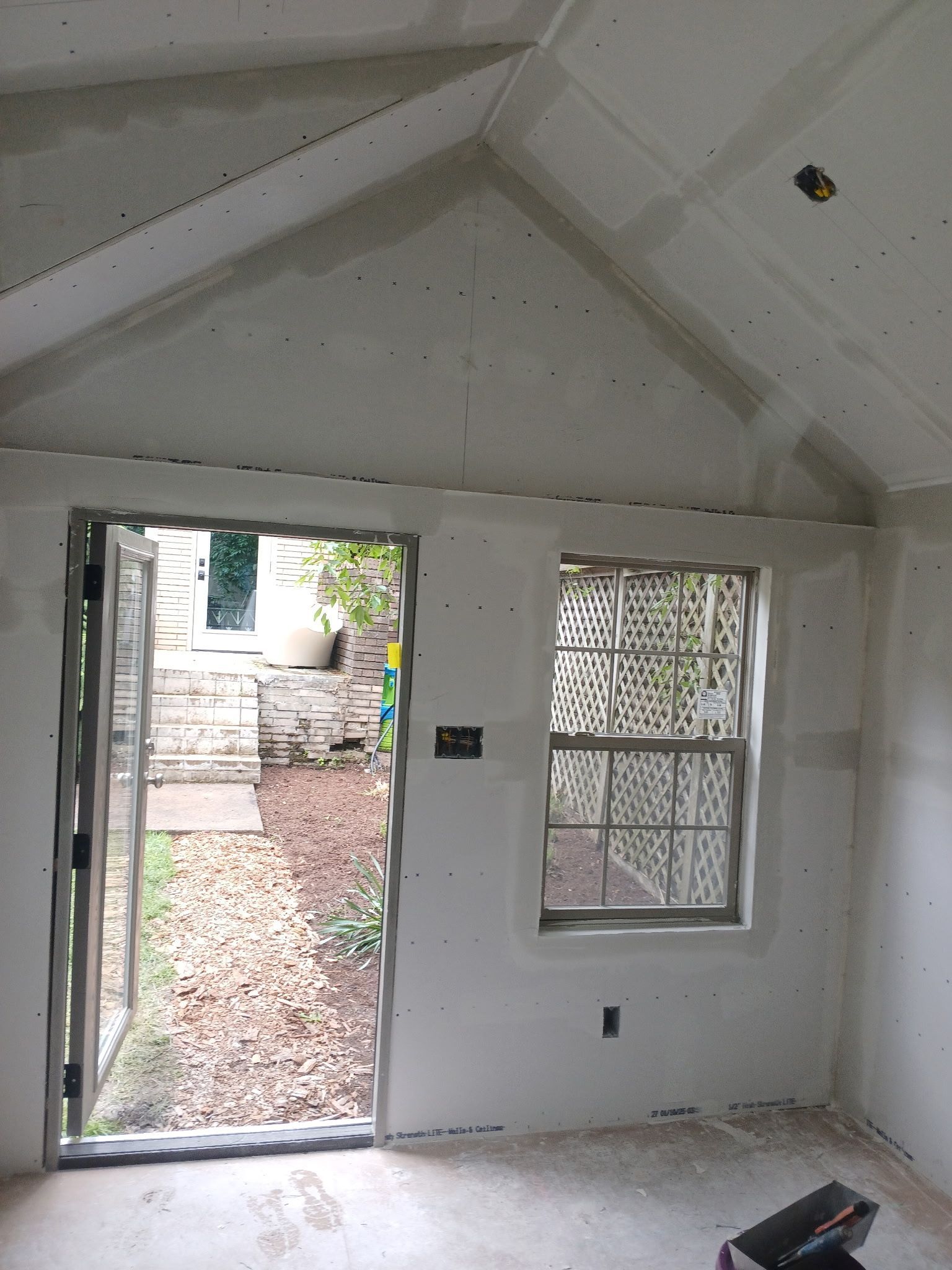 Empty unfinished room with open doorway and window, showing a sloped ceiling and outdoor path.