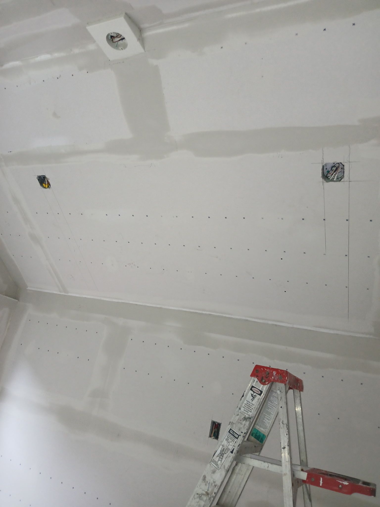 Ceiling drywall under construction with taped seams, screw patches, and a ladder below