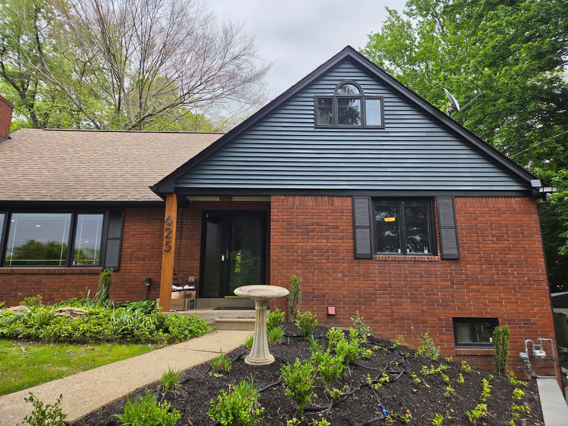 Brick house with dark roof, front walkway, and landscaped garden under green trees