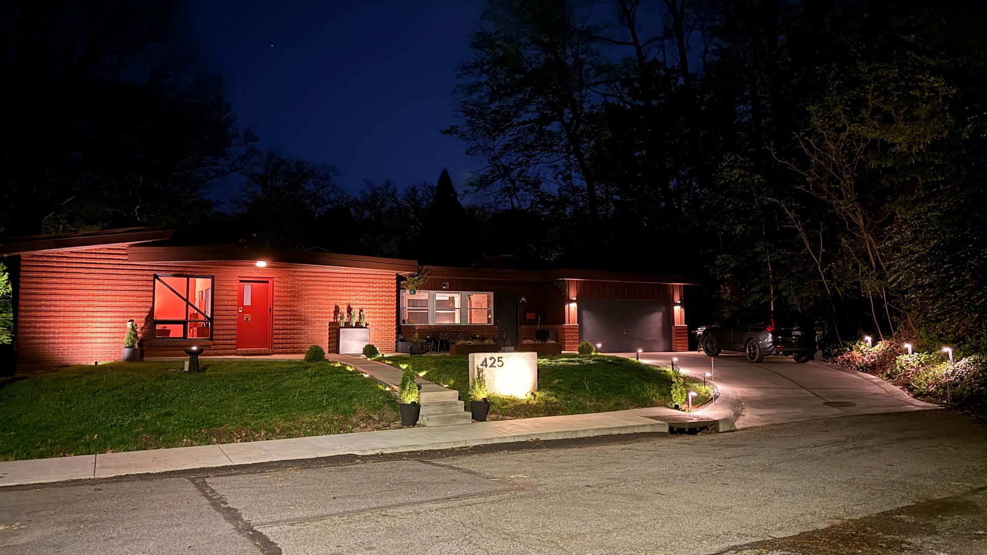 Single-story brick house lit warmly at night, with a lit walkway, driveway, and trees in front.