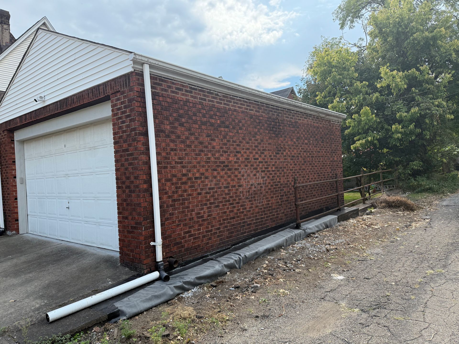 Brick garage with white door and downspout, beside a gravel driveway and trees under a cloudy sky