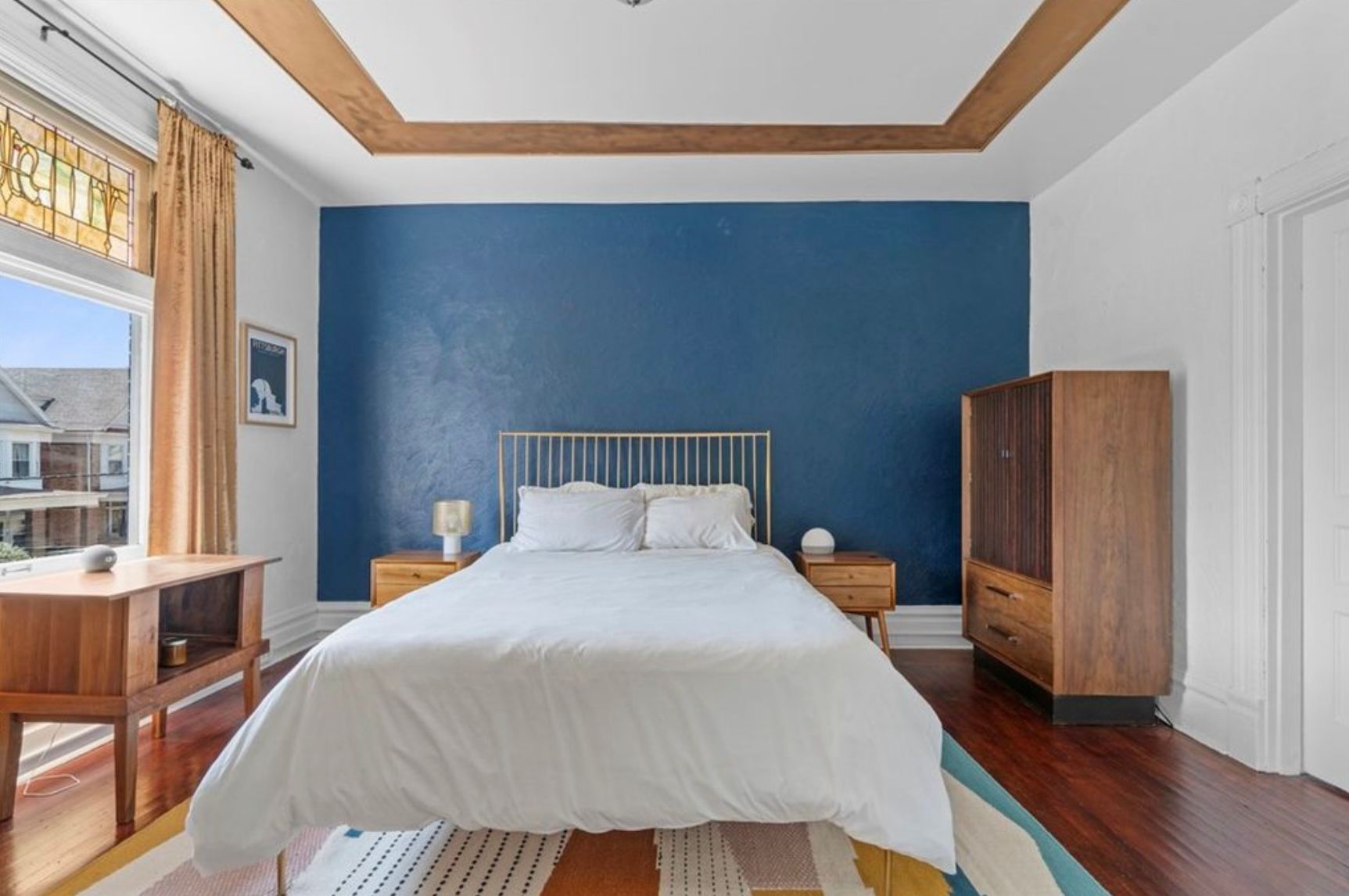 Bedroom with a blue accent wall, white bed, wooden dresser, and large window with yellow curtains.