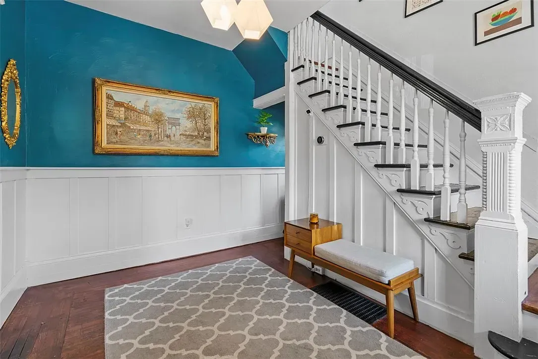 Blue foyer with white wainscoting, staircase, bench, patterned rug, and framed wall art