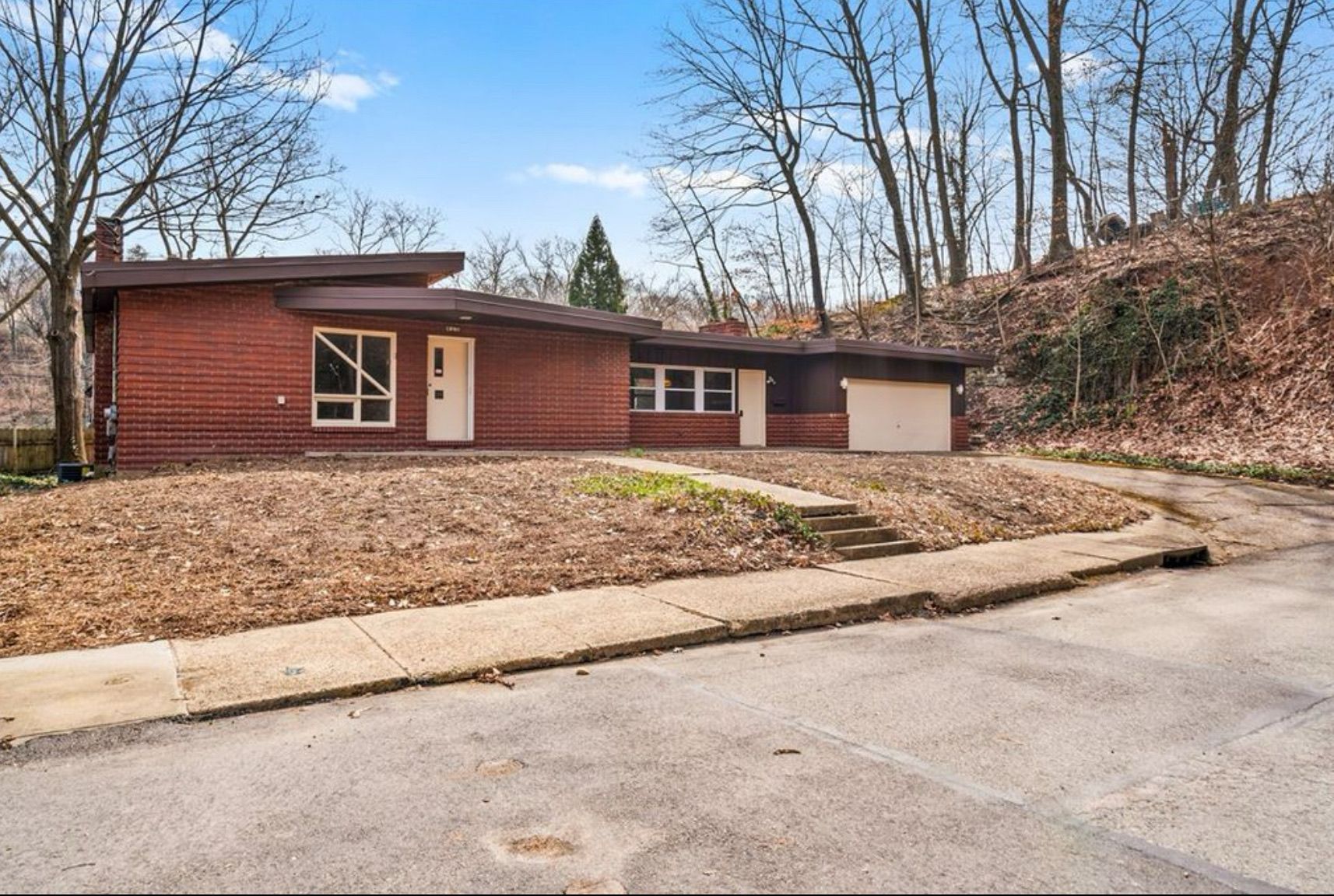Single-story brick house with a sloped front yard, garage, and leafless trees beside a road.