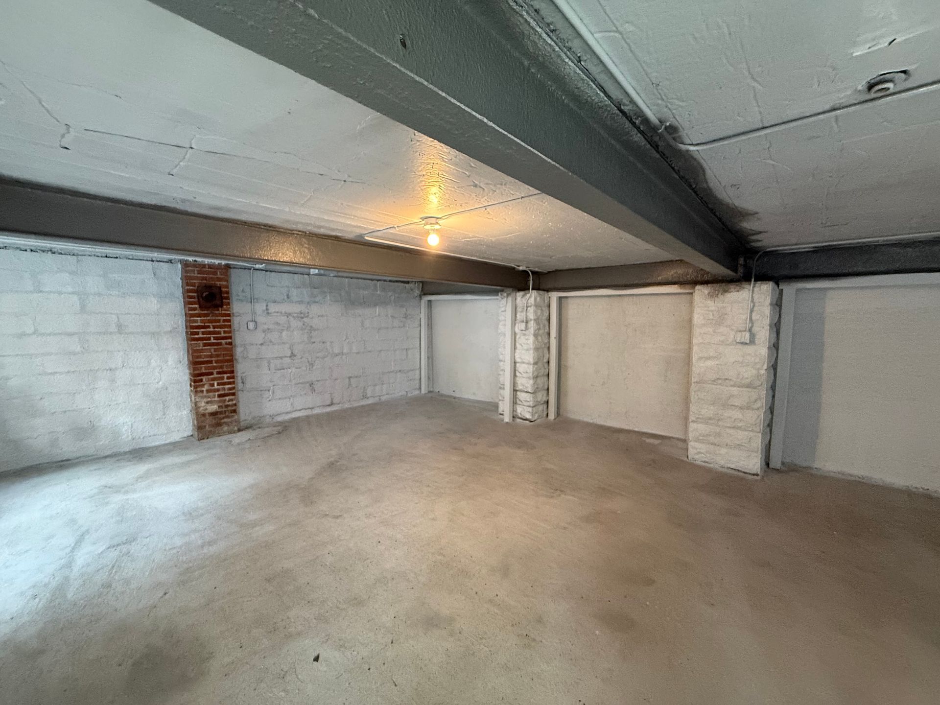 Empty concrete basement with white brick walls, support columns, and overhead beams under dim lighting