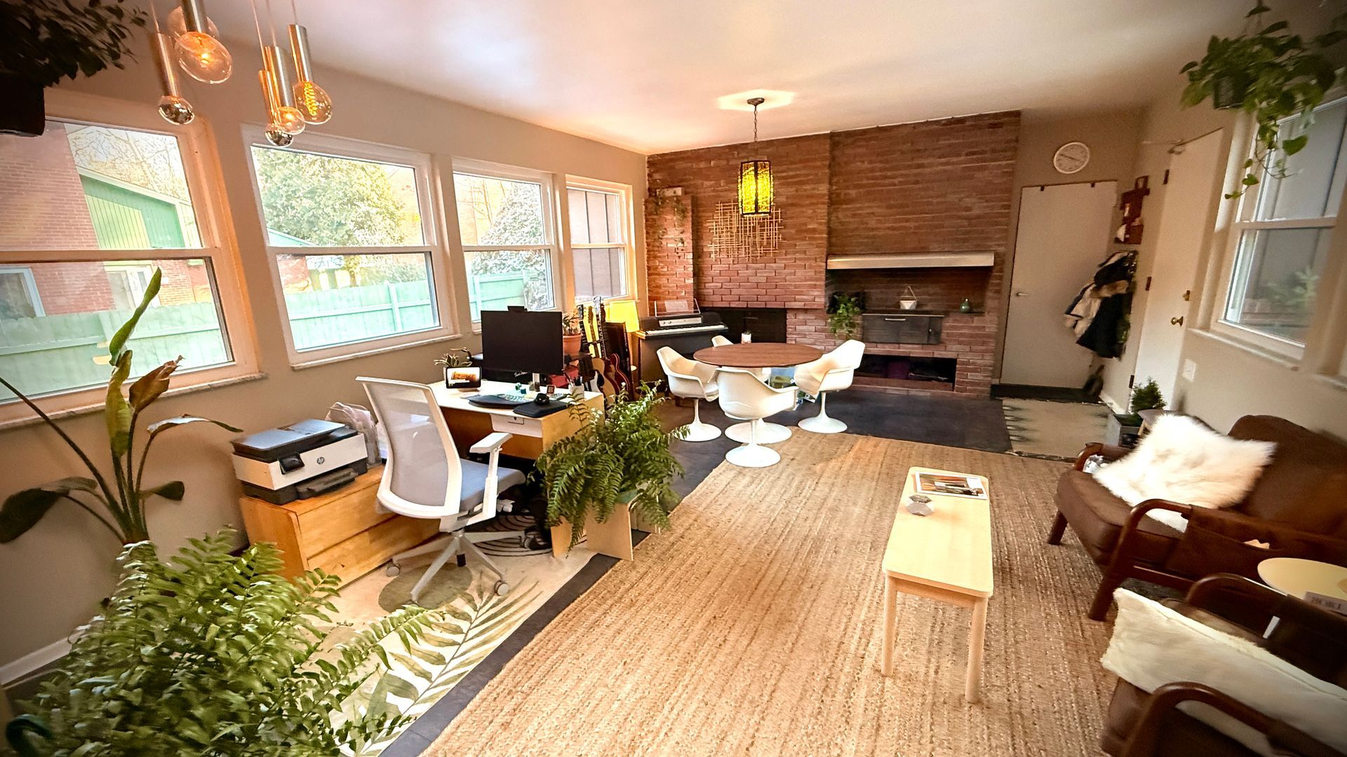 Bright living room with large windows, brick fireplace, woven rug, and modern chairs around a dining table.