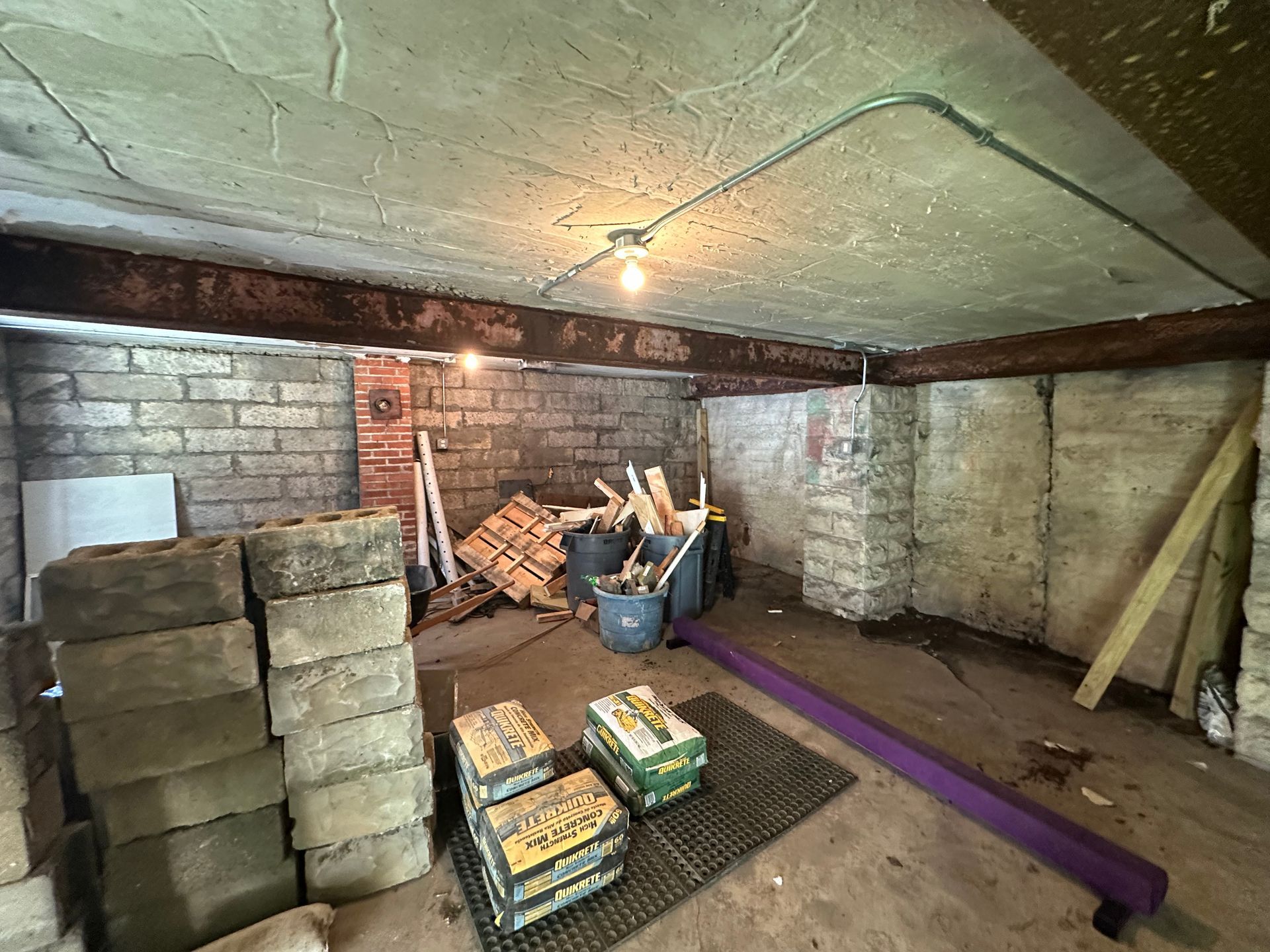 Dim basement storage room with stacked blocks, tools, a purple beam, and exposed brick walls under a low ceiling