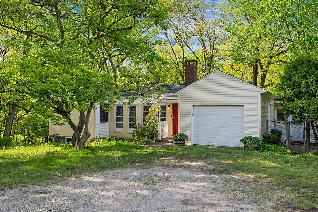Small white house with red door, garage, and gravel driveway surrounded by trees in a leafy yard