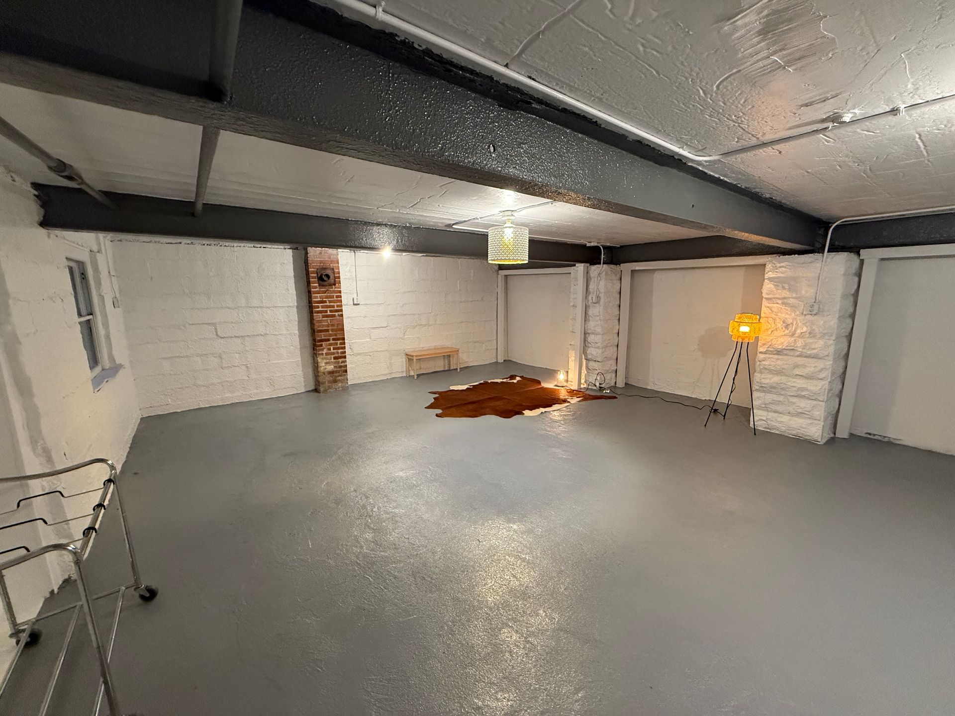 Empty basement with white brick walls, gray floor, exposed beams, and a small orange pile near the back wall