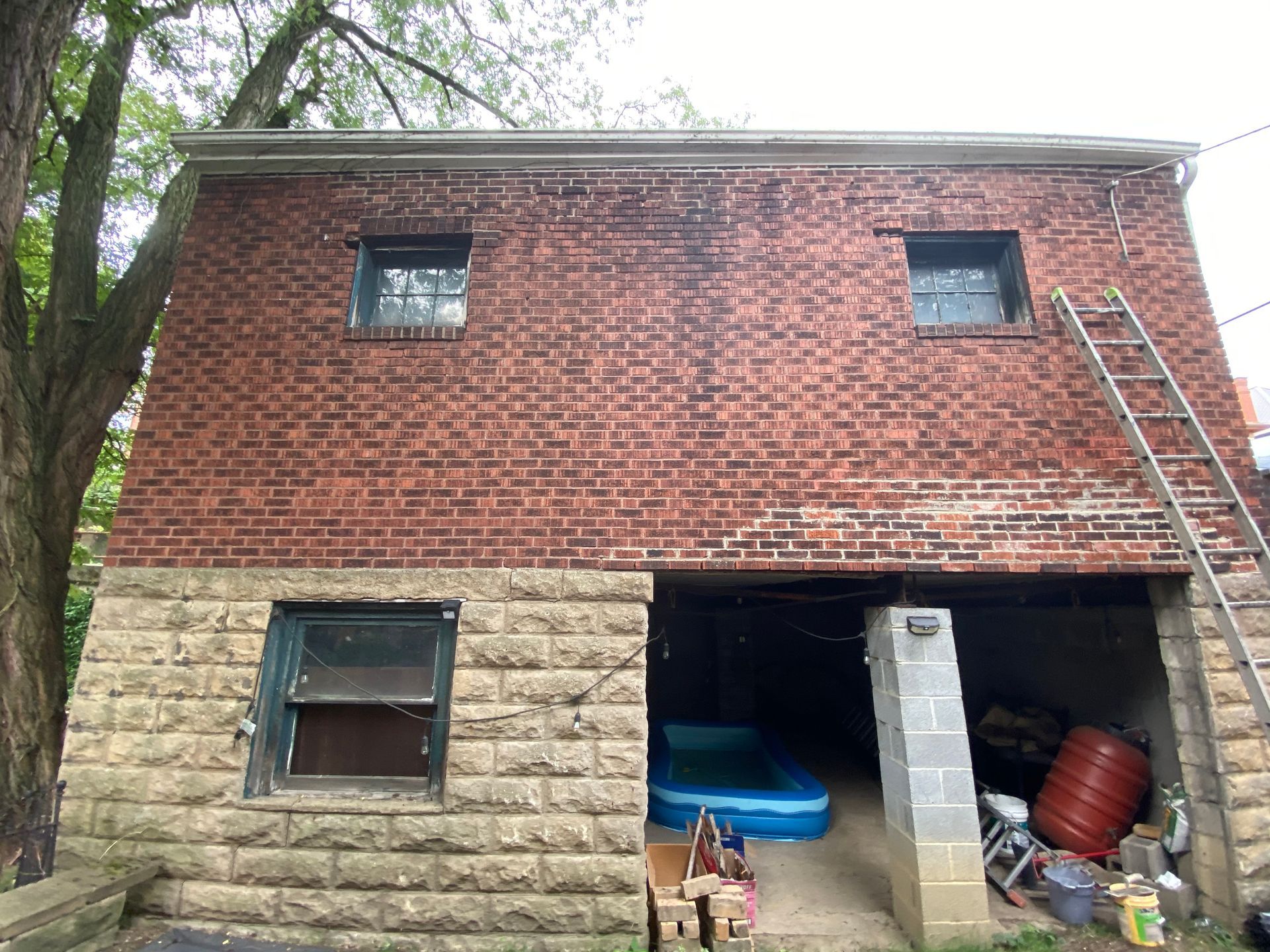 Red brick and stone house exterior with open garage, ladder, and blue pool visible inside