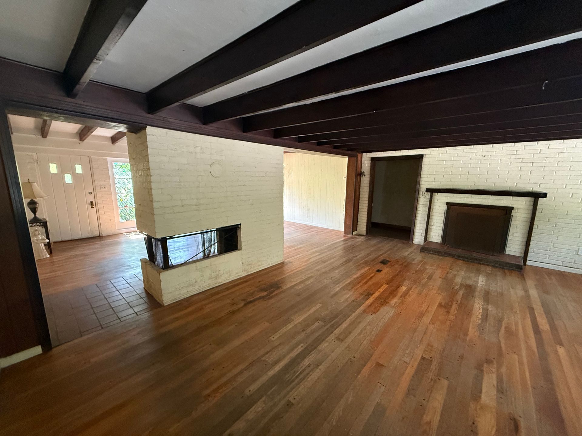 Empty living room with hardwood floors, exposed dark ceiling beams, and a fireplace on the right.