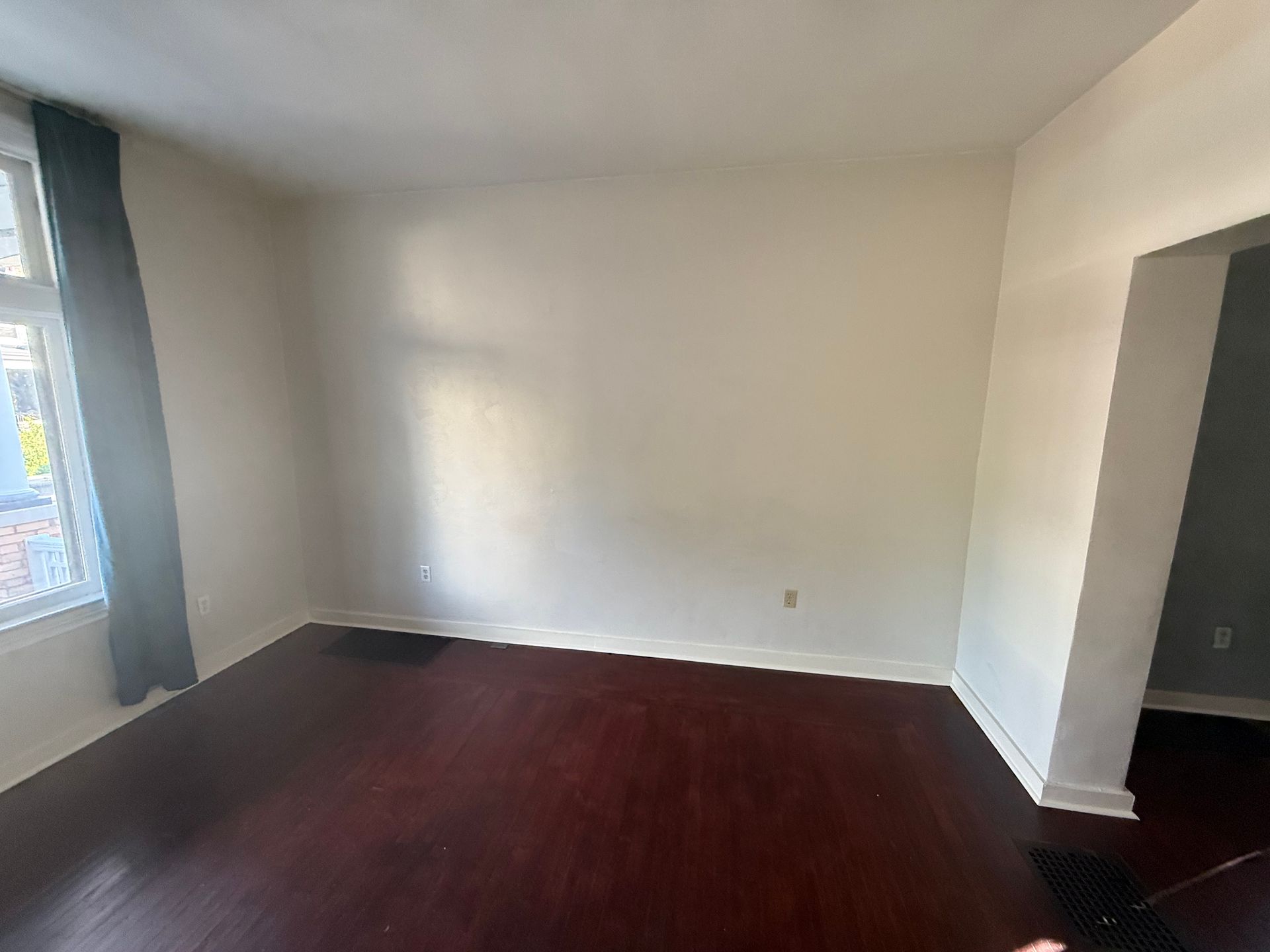 Empty living room with dark wood floors, white walls, and a window with dark curtains.